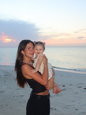 Una mujer de cabello largo oscuro sonríe mientras carga a una niña pequeña. Detrás se ven el mar y el cielo con tonos rosados al atardecer