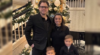 Pareja sonriente de adultos frente a un árbol de Navidad decorado y una escalera. Dos niños borrosos en primer plano, piano de cola al fondo.