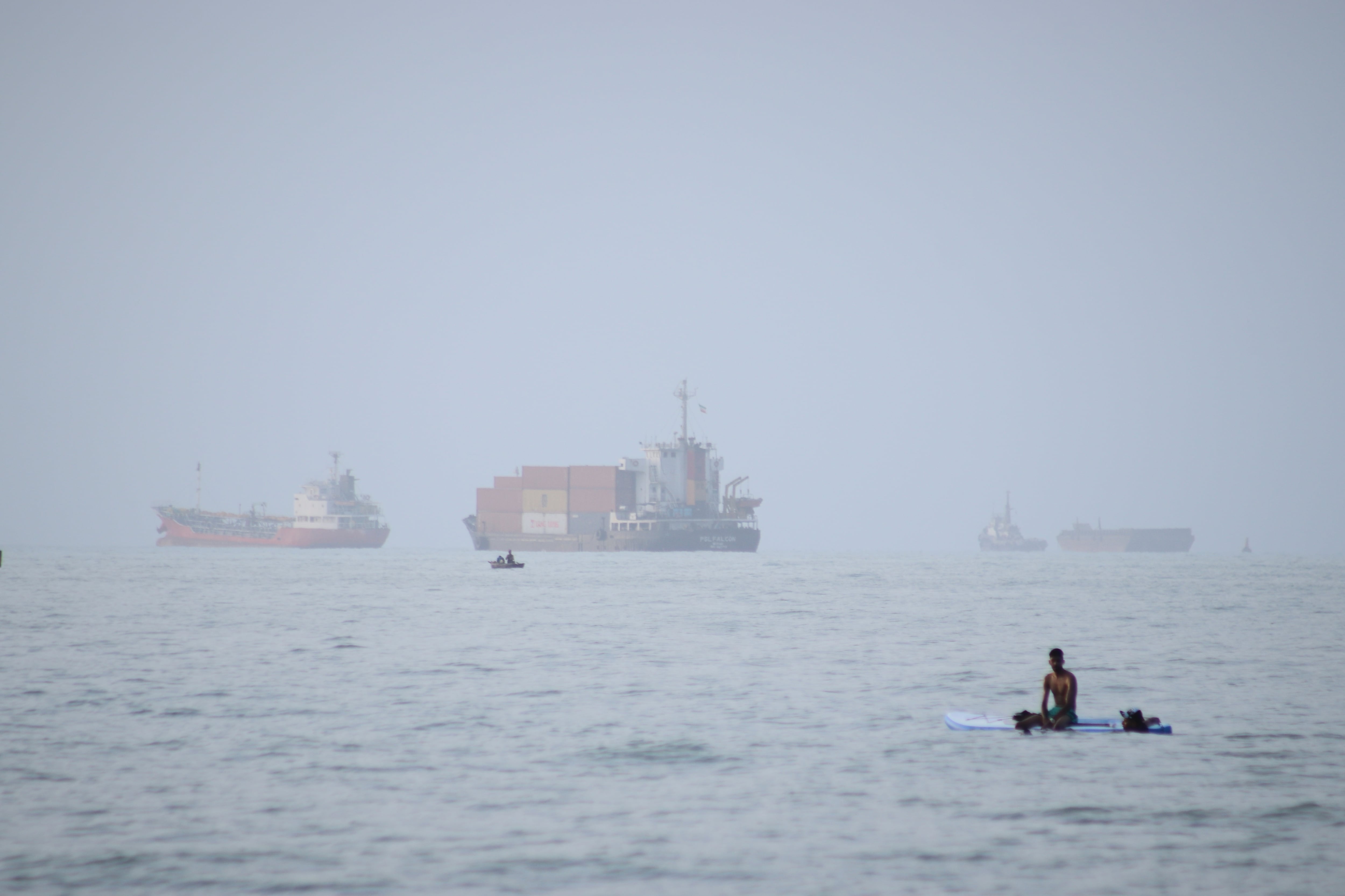 ARCHIVO - Un hombre se sienta en una tabla de surf con un barco portacontenedores al fondo junto a otros buques comerciales, en el estrecho de Ormuz, desde Bandar Abbas, irán, el lunes 27 de julio de 2026. (Razieh Poudat/ISNA via AP, Archivo)