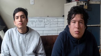 Dos hombres jóvenes están sentados ante una pizarra con inscripciones y un archivador gris de metal en una habitación.