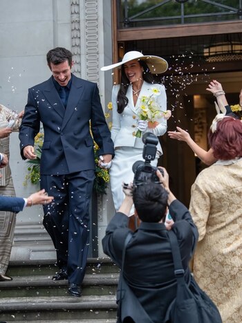 Una pareja camina por unas escaleras, con confeti cayendo sobre ellos. Ella lleva un traje blanco y sombrero. Él viste un traje oscuro. Un fotógrafo en primer plano