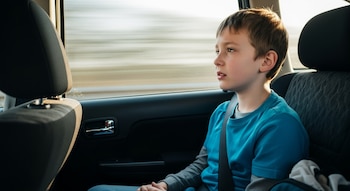 Niño en el asiento trasero de un automóvil con cinturón de seguridad, mirando por la ventanilla hacia un paisaje borroso con luz natural.