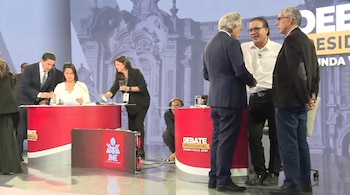 Personas en un escenario televisivo; dos hombres conversan, mientras otros revisan documentos en una mesa roja. El fondo muestra una imagen arquitectónica.