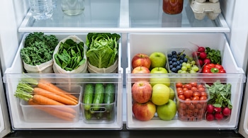 Cajones transparentes de un refrigerador con lechugas, zanahorias, pepinos, manzanas, uvas, jitomates y rábanos en recipientes.