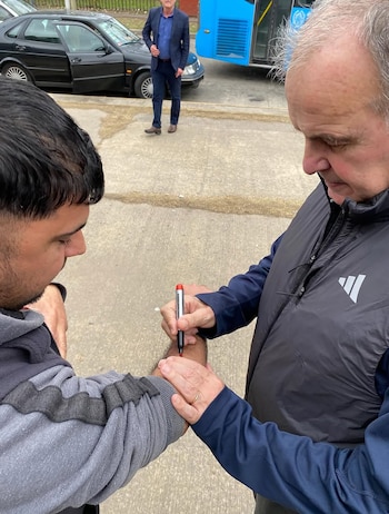 Un hombre de cabello gris firma con un marcador el brazo de otro hombre joven. Al fondo, un coche negro con la puerta abierta, un autobús azul y un hombre en traje