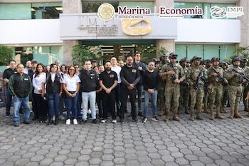Un grupo de personas vestidas de civil y varios militares uniformados con armas posan frente a la fachada de un edificio del IMPI en México