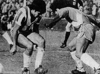 Fotografía en blanco y negro de dos futbolistas en un campo de juego disputando una pelota frente a las tribunas