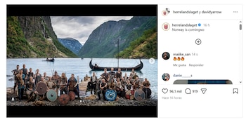 Un grupo de hombres vestidos de vikingos con escudos y armas posa a orillas de un fiordo noruego, con montañas y un drakkar en el fondo