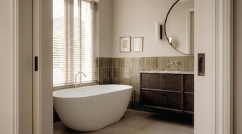 Vista de un baño moderno con bañera blanca, mueble de madera oscura, azulejos verdes, espejo redondo y ventana con persianas.