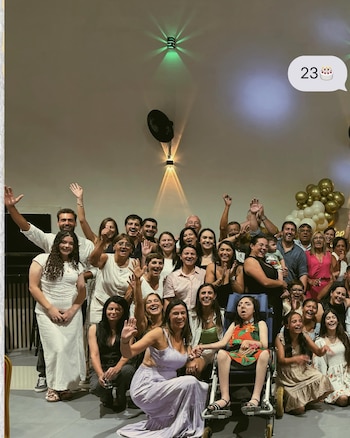 Gran grupo de personas sonriendo y agitando las manos en un salón. Una joven en silla de ruedas con vestido estampado se ubica en el centro