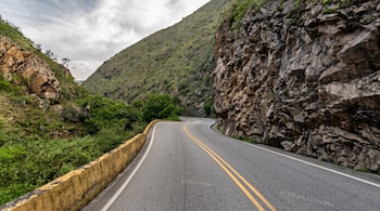 Carretera pavimentada con doble línea amarilla, bordeada por una pared de roca a la derecha y vegetación con un murete a la izquierda.