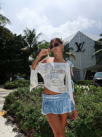 Mujer con cabello recogido, gafas de sol, blusa con estampado floral, falda azul de terciopelo. Palmeras, arbustos y tienda Louis Vuitton de fondo