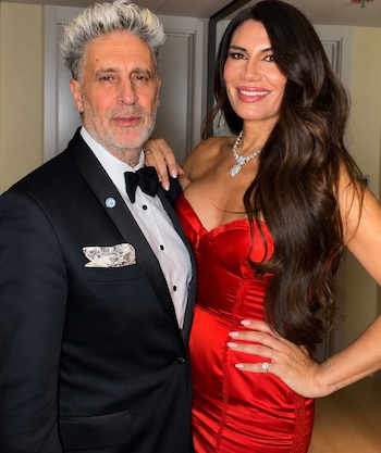 Hombre de pelo y barba canosos con esmoquin negro, camisa blanca, moño negro y pin en la solapa posa junto a mujer de cabello oscuro con vestido rojo strapless y joyas