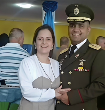 Un hombre con uniforme militar verde oscuro y gorra sonríe junto a una mujer con blusa blanca y top beige, tomada de su mano. Al fondo se aprecian otras personas