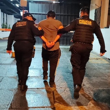 Vista trasera de dos policías con uniformes negros y chalecos tácticos escoltando a un hombre esposado por la espalda. Caminan sobre un pavimento mojado