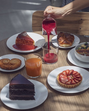 Una mano vierte una bebida roja en un vaso sobre una mesa de madera con un surtido de pasteles, tartas, bollería y zumo de naranja