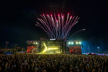 Vista general de un multitudinario festival de música nocturno con fuegos artificiales rojos y blancos explotando sobre un escenario muy iluminado