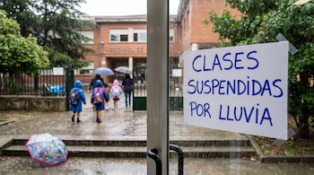 Un cartel pegado en una puerta de vidrio indica 'CLASES SUSPENDIDAS POR LLUVIA' frente a niños con paraguas en un patio mojado.