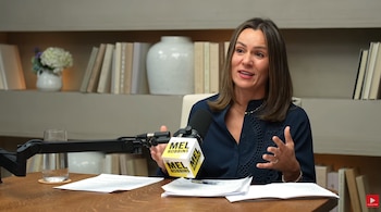 Una mujer sentada a una mesa de madera con hojas impresas, un vaso de agua y un micrófono con el logotipo de The Mel Robbins Podcast