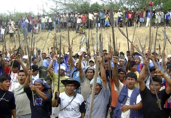 Una multitud de hombres indígenas, muchos con pintura facial y vestimenta tradicional, sostienen lanzas de madera en alto, gritando en señal de protesta