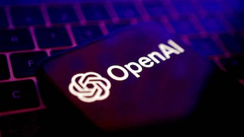 Primer plano de una pantalla de smartphone oscura con el logo blanco y el texto 'OpenAI' sobre un teclado de ordenador con poca luz