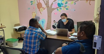 Una mujer con mascarilla sentada en un escritorio frente a dos hombres. La pared rosa tiene un colorido mural de un árbol con monos, búhos y cebras