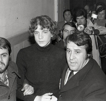 Joven con cabello rizado y suéter de cuello alto es escoltado por un hombre con traje, rodeados por una multitud y fotógrafos con cámaras y flashes