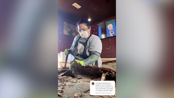 Mujer con mascarilla, gafas verdes y guantes, usando una palanca para retirar trozos de madera de un suelo, con un televisor mostrando un dibujo animado al fondo
