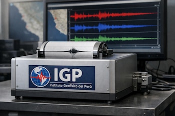 Sismógrafo del Instituto Geofísico del Perú sobre una mesa, con un registro de ondas sísmicas. Un monitor muestra ondas sísmicas en colores y mapas desenfocados.
