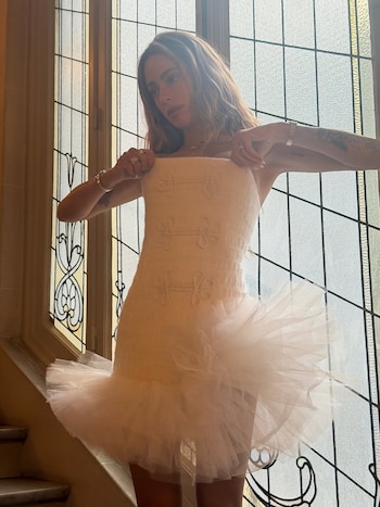 Mujer con cabello claro y tatuajes, vestida con un minivestido blanco sin tirantes de cuerpo texturizado y falda de tul. Está frente a una ventana con paneles
