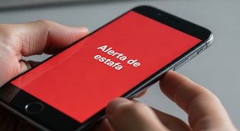 Primer plano de unas manos sosteniendo un teléfono celular moderno con una pantalla roja brillante que muestra el texto en blanco "Alerta de estafa".