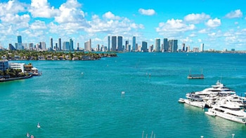 Una vista aérea de la ciudad de Miami con numerosos rascacielos al fondo, el océano azul vibrante en primer plano con varios yates blancos y pequeñas embarcaciones
