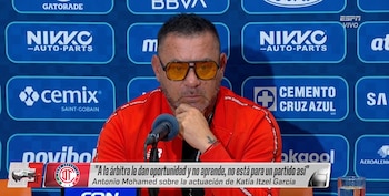 Antonio Mohamed habla frente a un micrófono con gafas amarillas y chaqueta roja ante un panel con logotipos publicitarios