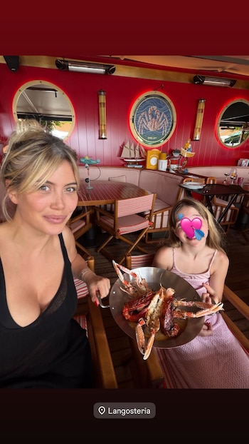 Wanda Nara sonríe en primer plano junto a una niña sentada que sostiene un plato de langostinos y mariscos en un restaurante de paredes rojas