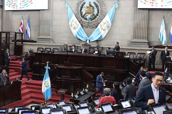 Un salón parlamentario con diputados sentados en escritorios y de pie. Banderas de Guatemala, Costa Rica y Honduras, y el escudo nacional de Guatemala decoran la sala