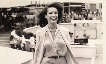 Fotografía en blanco y negro de una mujer sonriente con cabello oscuro y voluminoso, vestida con un blazer y pantalón claro, con personas y edificios al fondo