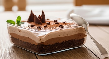 Postre de brownie sin cocción en un recipiente de vidrio, con capas de chocolate, base de galleta, decorado con cacao, virutas y menta. Cuchara en mesa de madera.