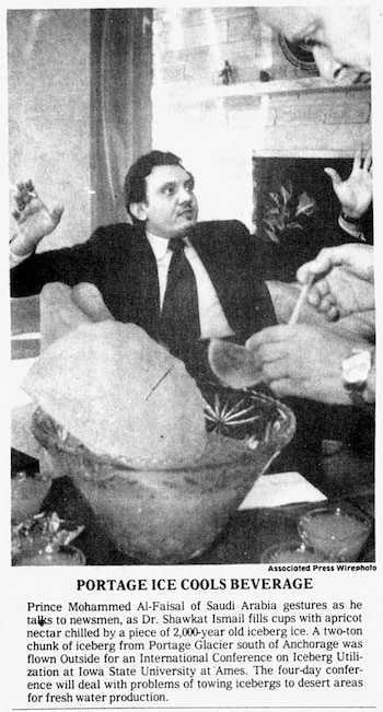 Hombre en traje gesticulando, sentado frente a una mesa con un gran trozo de hielo en un cuenco. Manos sirviendo una bebida en un vaso