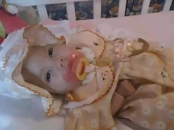 Primer plano de un bebé o muñeca en una cuna, con un gorro de tela claro, un chupete rosa y amarillo en la boca y un tubo discreto cerca de la nariz