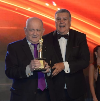 Dos hombres con traje de etiqueta posan en un escenario iluminado mientras uno de ellos sostiene un premio dorado