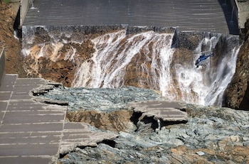 Un helicóptero vuela junto al agua que cae por una ladera rocosa entre losas de concreto fracturadas de una represa