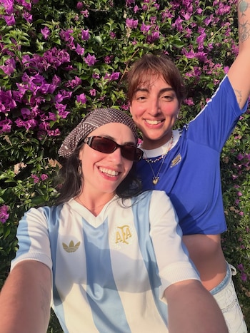Dos personas sonrientes, hombre y mujer, frente a un arbusto de flores púrpuras. La mujer usa pañuelo y gafas de sol. Ambos visten camisetas de fútbol celestes y blancas