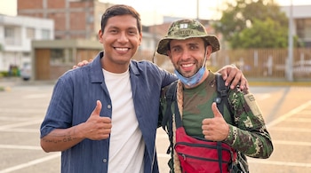 Dos hombres sonrientes con ropa informal posan al aire libre haciendo un gesto con el pulgar hacia arriba. Uno lleva una camisa azul, el otro uniforme de camuflaje y gorra.