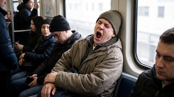 Hombre con gorro verde bosteza en un tren. Otros pasajeros están sentados o de pie en el vagón.