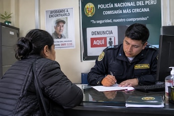 Un agente de policía escribe en un documento frente a una mujer en un escritorio, con carteles institucionales y de búsqueda al fondo