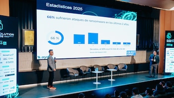Un hombre de traje y zapatillas rojas presenta estadísticas de ransomware en una pantalla grande a una audiencia en un escenario con sillas y mesas, con otro hombre al costado
