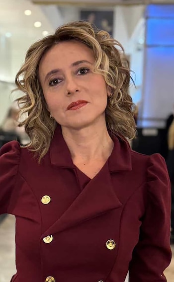 Primer plano de una mujer con cabello rizado rubio, blazer borgoña de doble botonadura con cuatro botones dorados, labios rojizos