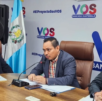 Hombre de tez morena habla en un micrófono en una mesa de madera. Detrás hay una bandera de Guatemala y logos del partido VOS