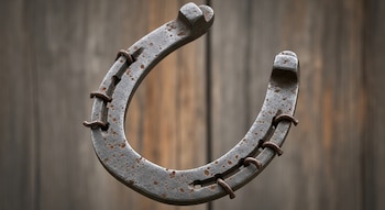 Una herradura de metal oxidado con seis clavos se muestra en primer plano, con un fondo desenfocado de tablones de madera con tonos marrones y grises.