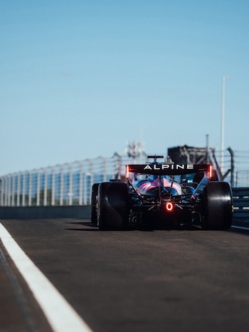 Vista trasera de un coche Alpine de Fórmula 1 en una pista de carreras, con luces rojas traseras, neumáticos anchos y cielo azul